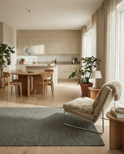 Modern beige kitchen and living area featuring a round wood dining table, wool rug, and a cozy armchair with a throw.