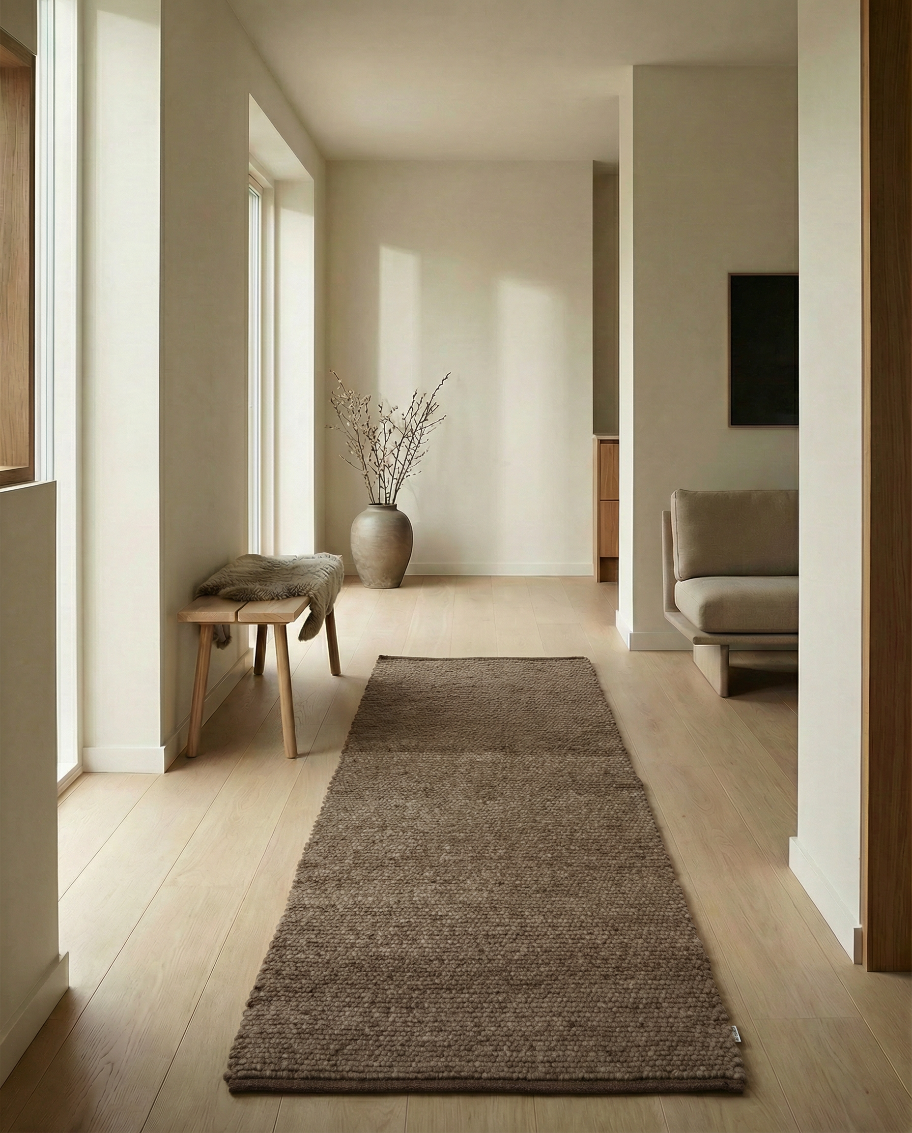 Minimalist hallway featuring a textured brown wool runner rug on a light oak floor with a wooden bench and vase.
