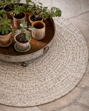 Rund ullmatta Merino i ull, naturbeige melerad, med cirkulär struktur, delvis täckt av lågt bord med krukväxter