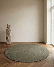 Large, circular olive green jute rug with a textured coiled pattern on a wooden herringbone floor.