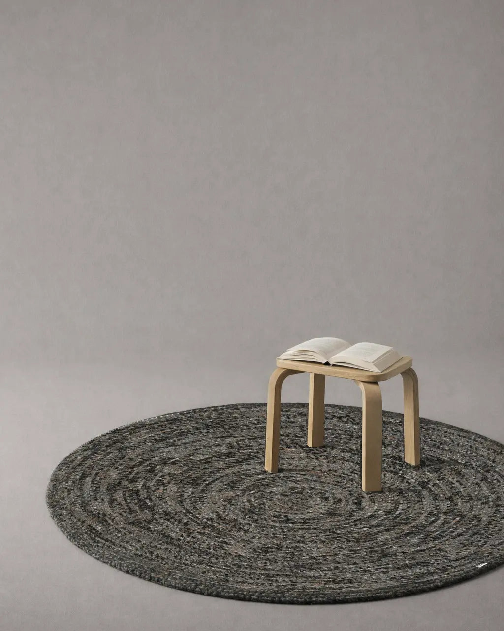Rund ullmatta Merino i grey melange placerad i avskalad interiör med pall och öppen bok