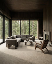 Modernes Wohnzimmer mit grauem Ecksofa, Steincouchtisch und Sessel vor bodentiefen Fenstern mit Waldausblick.