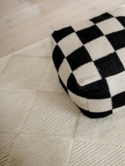 Puff Square Svart/Natur, 50x50 cm, handvävd i ull, placerad på en ljus strukturerad ullmatta.