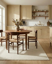 Round dark wood dining table with woven-seat chairs in a modern, light beige kitchen with shaker cabinets.