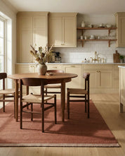 Round wooden dining table with chairs featuring woven seats on a terracotta rug in a beige modern kitchen.