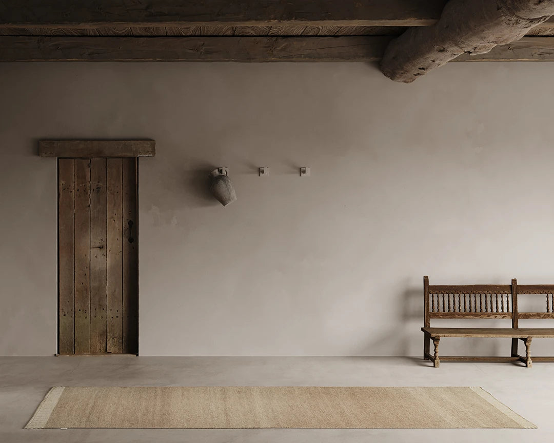 Gångmatta Pure Natural Beige i ull, 80x250 cm, i avlång form på ljus betonggolv i rustik interiör med träbjälkar, gammal trädörr och träbänk.
