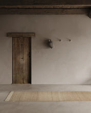 Gångmatta Pure Natural Beige i ull, 80x250 cm, placerad på ljus stenliknande golv i rustik hallmiljö med träbjälkar och gammal trädörr.