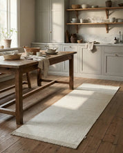 Off-white rectangular woven kitchen runner rug with fringe edges on rustic wooden floor in a farmhouse kitchen.