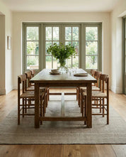 Rustikaler Esstisch aus Holz mit sechs Stühlen mit Geflechtsitz vor einer großen Fensterfront im hellen Wohnzimmer.
