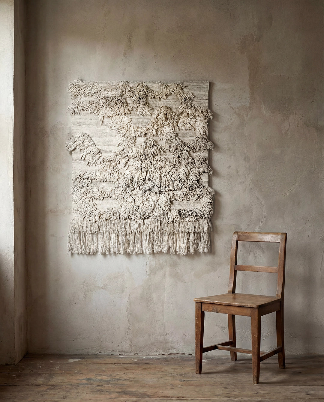Beige väggbonad i ull med strukturerad lugg och fransar, hängande på en vägg bredvid en trästol.