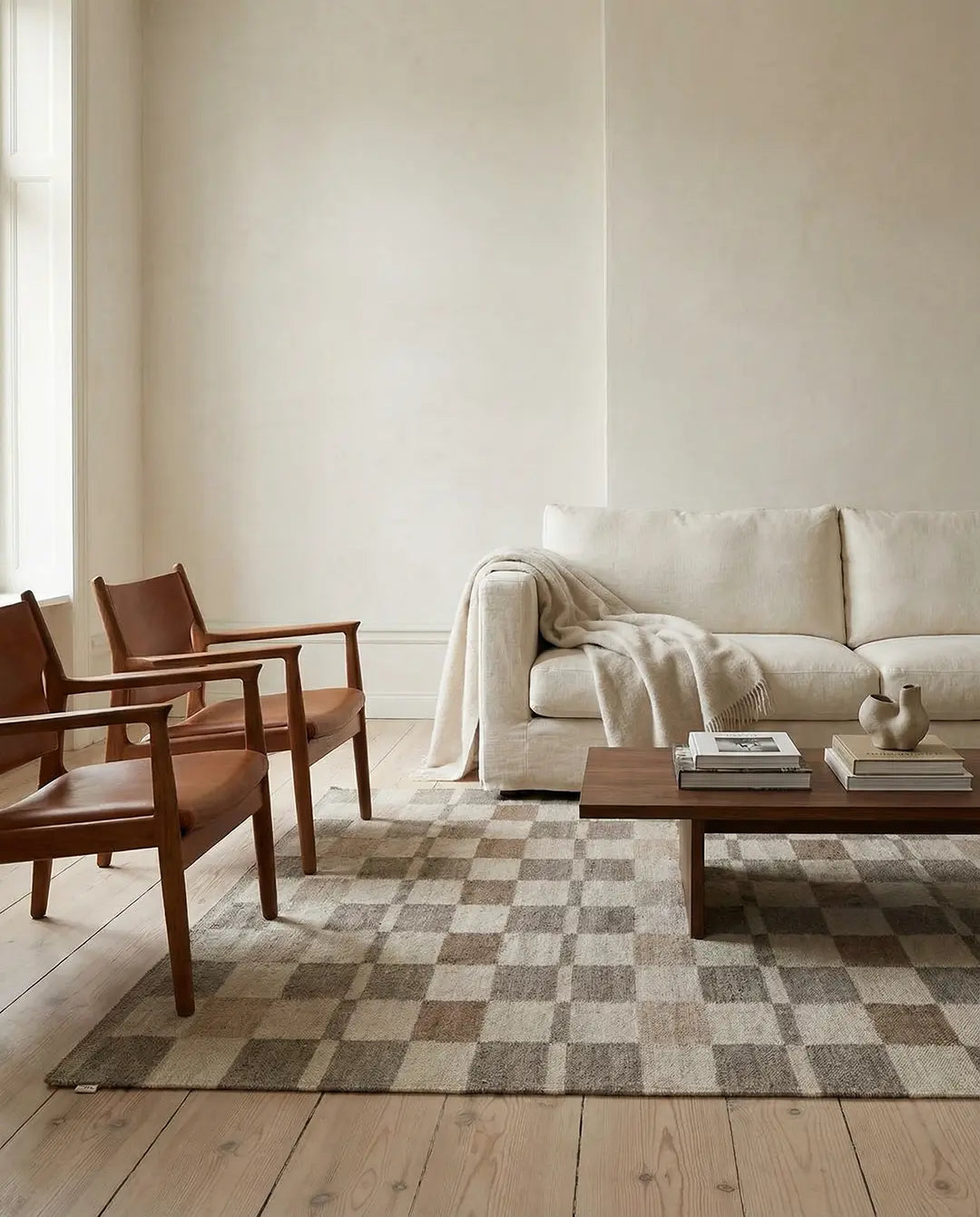 Beiges Sofa mit Decke, zwei braune Ledersessel und ein Teppich mit Schachbrettmuster in einem hellen Wohnzimmer.