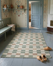 Rektangulär matta med rutigt mönster i dova nyanser av grönt, beige och creme på ett trägolv i lantlig miljö.