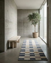 Moderner Flur mit schmalem Läufer in Blau-Beige Karo-Muster, Travertin-Bank und Olivenbaum vor Betonwänden.