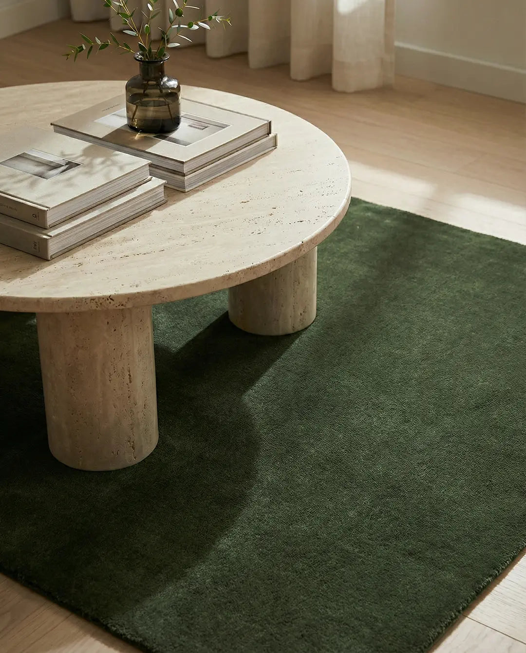 Runt soffbord i beige travertin på en mörkgrön matta i ett ljust vardagsrum.