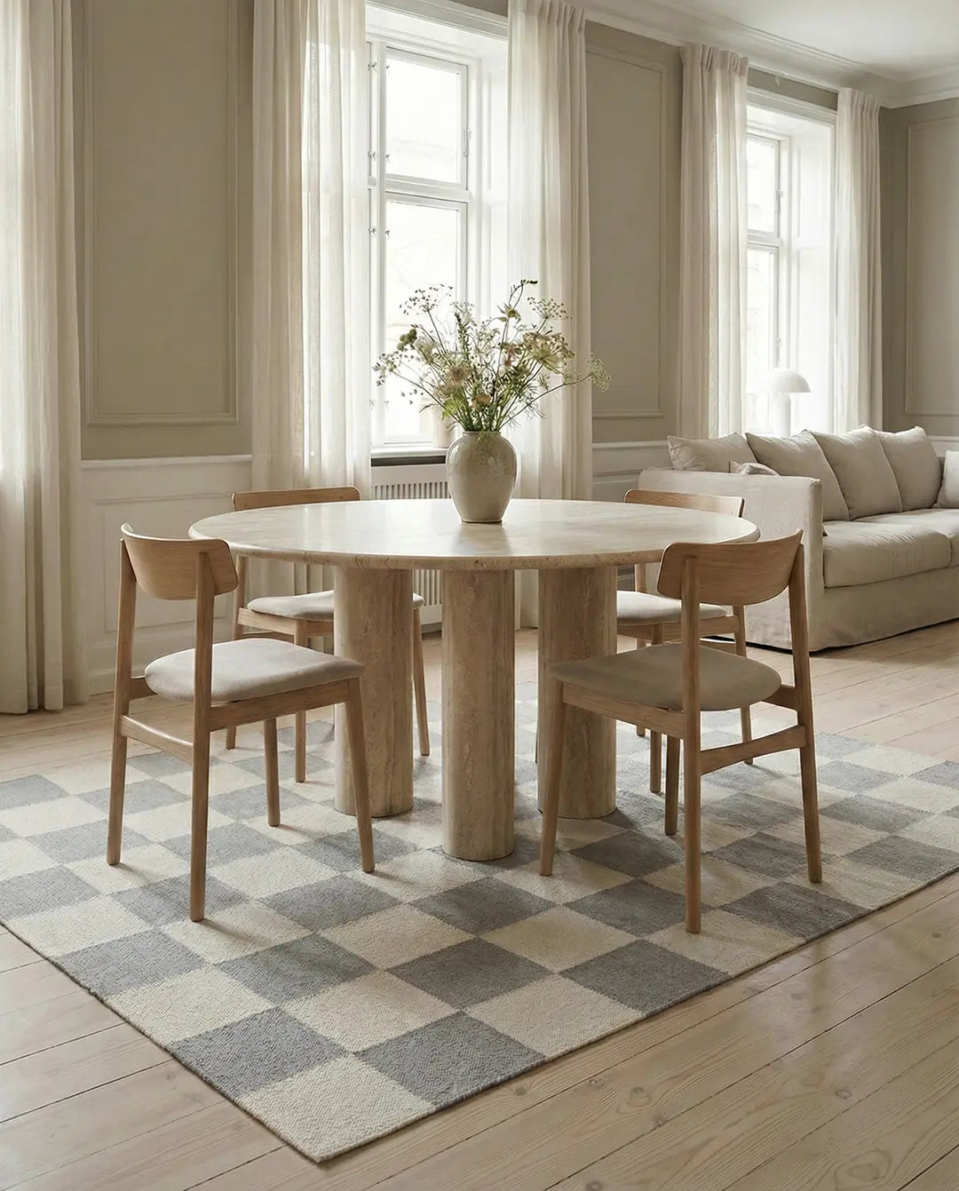 Runder Esstisch aus hellem Travertin mit Säulenfüßen und vier Holzstühlen auf einem Teppich mit Schachbrettmuster.