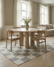 Runder Esstisch aus hellem Travertin mit Säulenfüßen und vier Holzstühlen auf einem Teppich mit Schachbrettmuster.