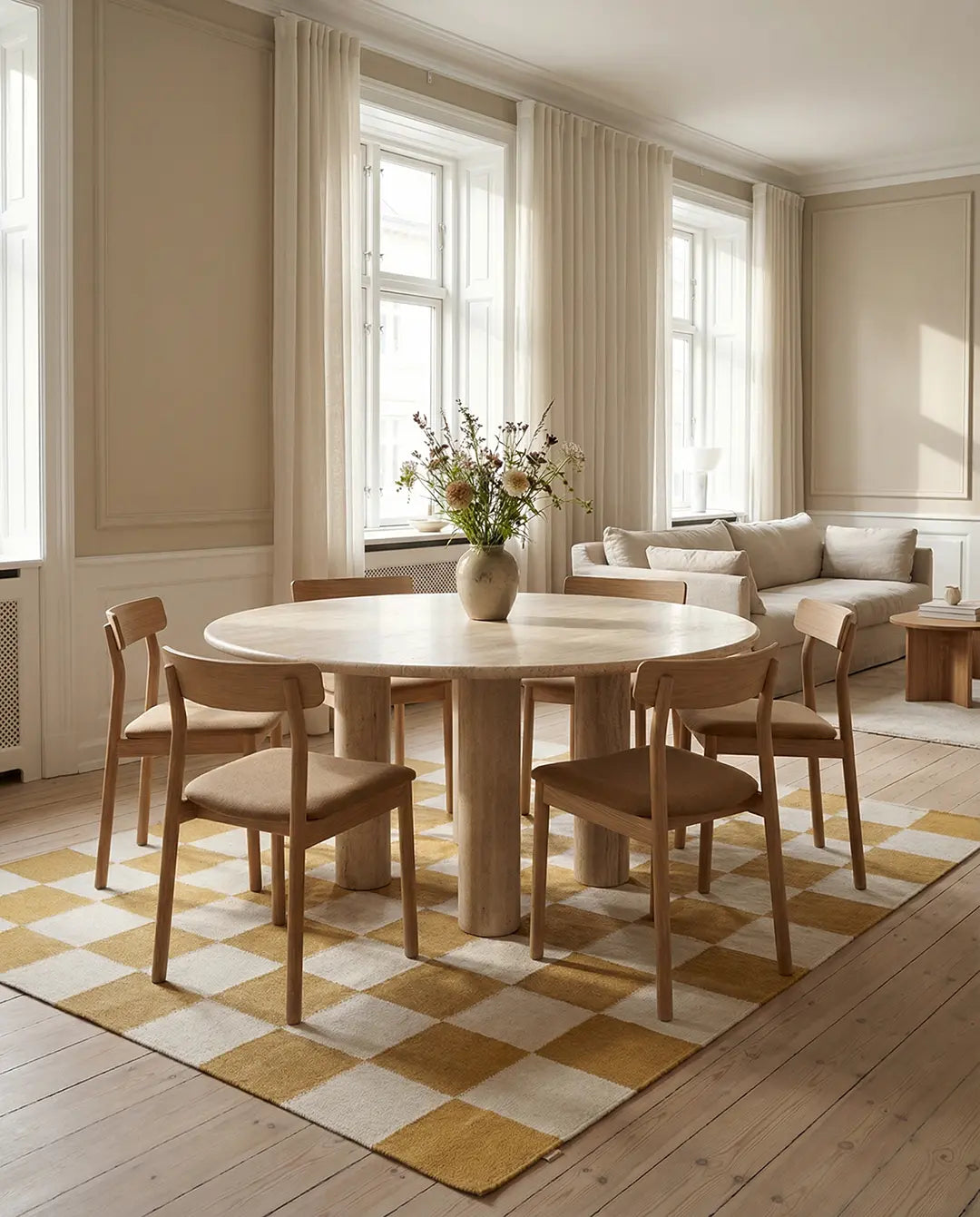 Runder Esstisch aus hellem Stein mit Säulenbeinen, umgeben von modernen Holzstühlen auf einem gelb-weißen Schachbrett-Teppich.