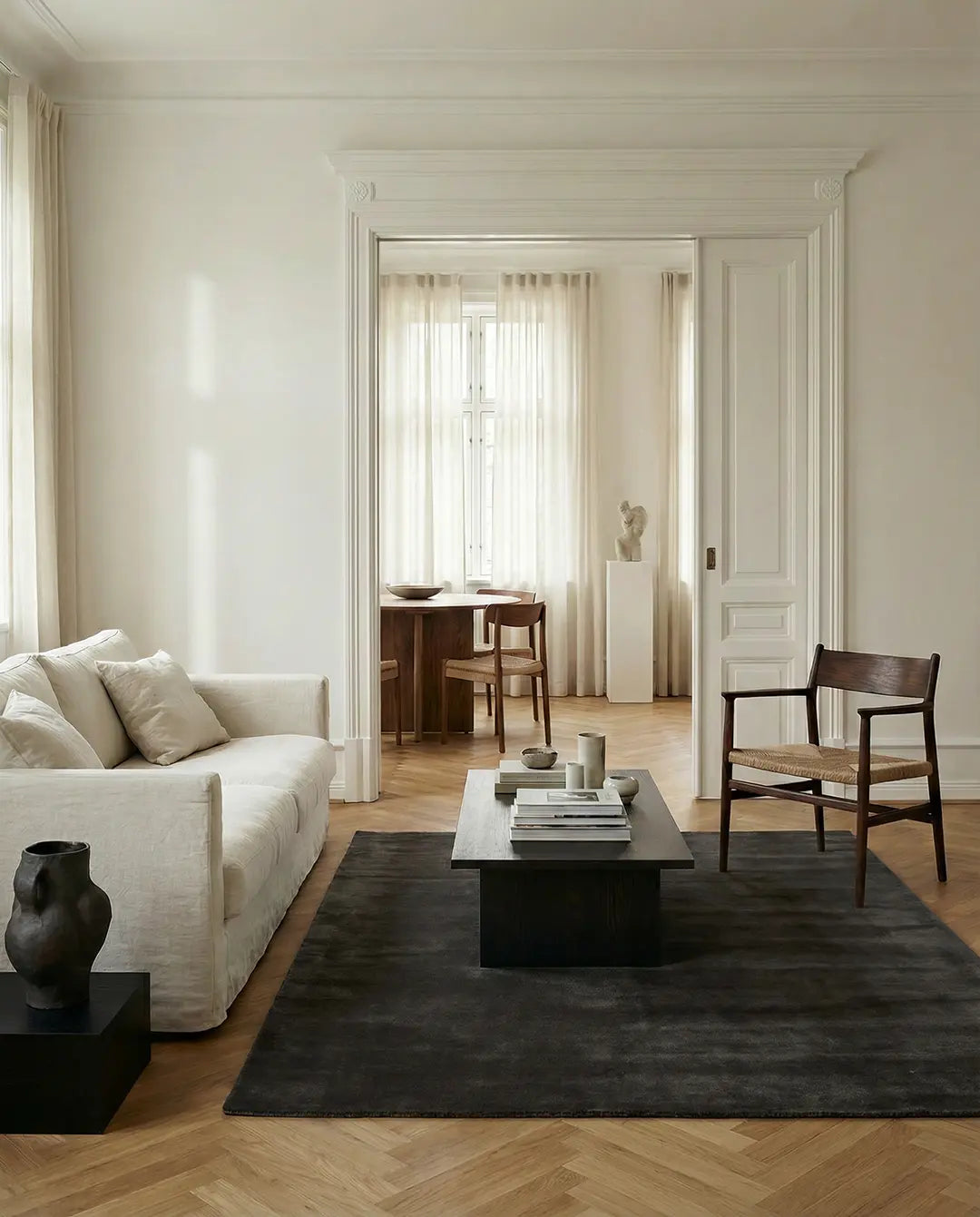 Ljust vardagsrum med beige soffa, mörkt träbord, fåtölj med flätad sits och mörkgrå matta i minimalistisk stil.