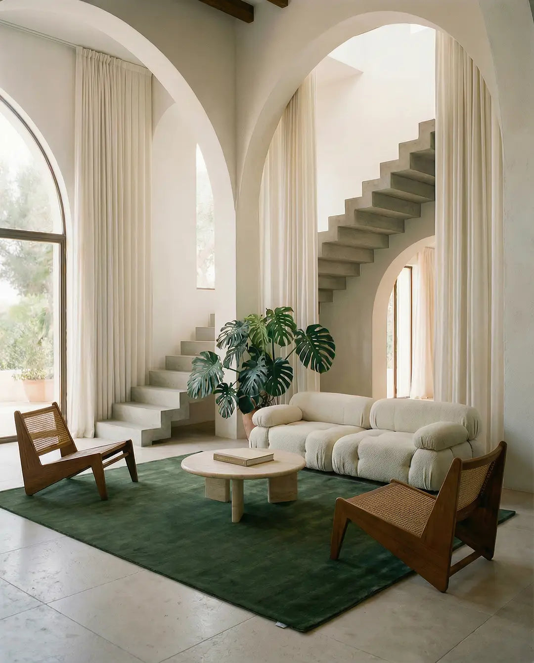 Modernt vardagsrum med beige soffa, rottingstolar i trä och en mörkgrön matta i ett ljust rum med välvda detaljer.