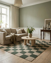 Wohnzimmer mit beigem Leinensofa, rundem Travertin-Couchtisch und grün-beigem Schachbrett-Teppich im modernen Landhausstil.