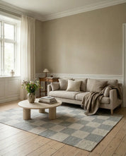 Beiger Couchtisch aus Holz mit runden Beinen auf einem grau-weißen, karierten Wohnzimmerteppich in einem hellen Raum.