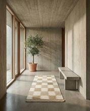 Rutig matta i beige och krämvitt på betonggolv i en minimalistisk hall med en olivträd och betongbänk.