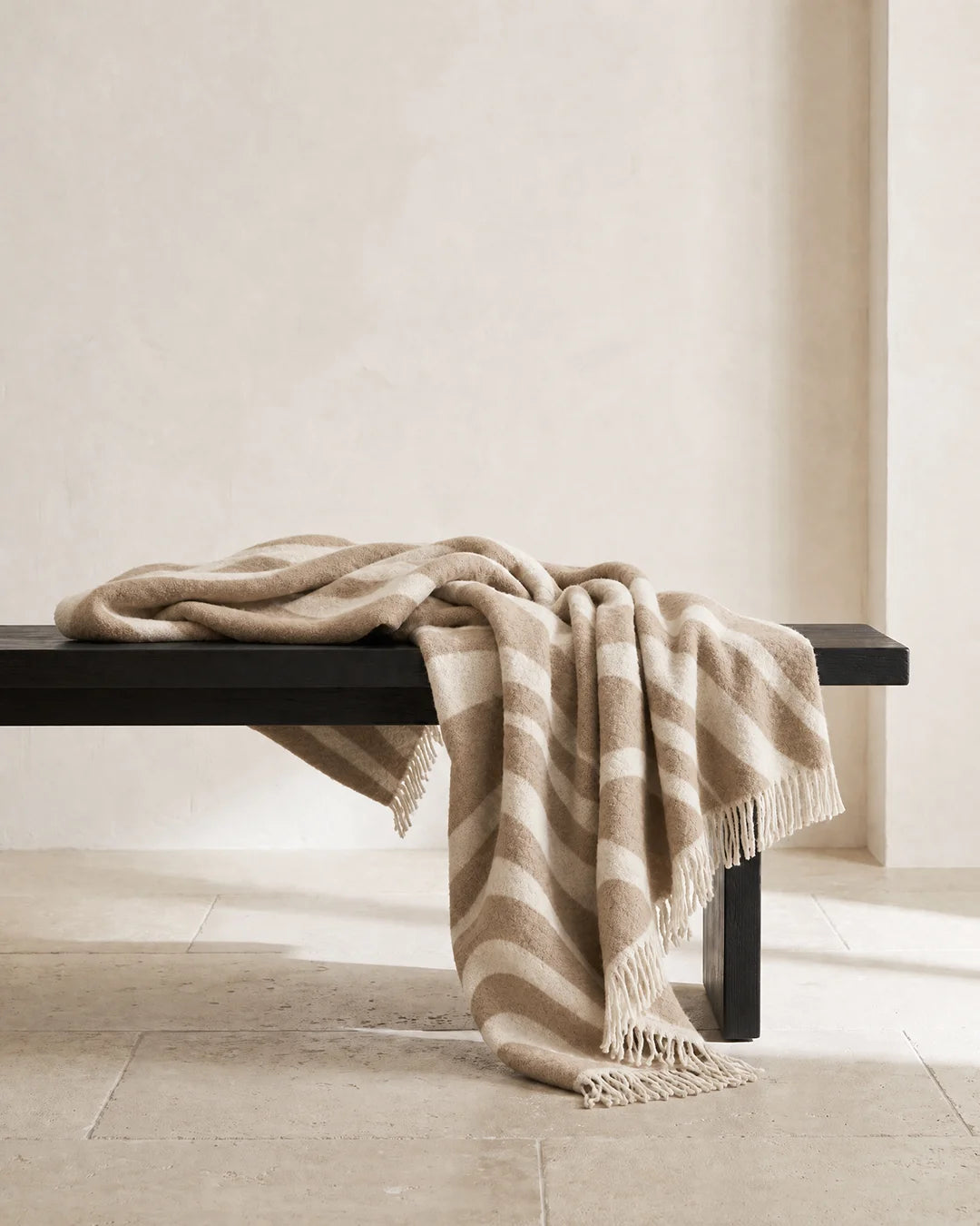 Pläd Flow Beige från Classic Collection i ull med vågformat mönster i beige och off-white, draperad över en mörk träbänk.