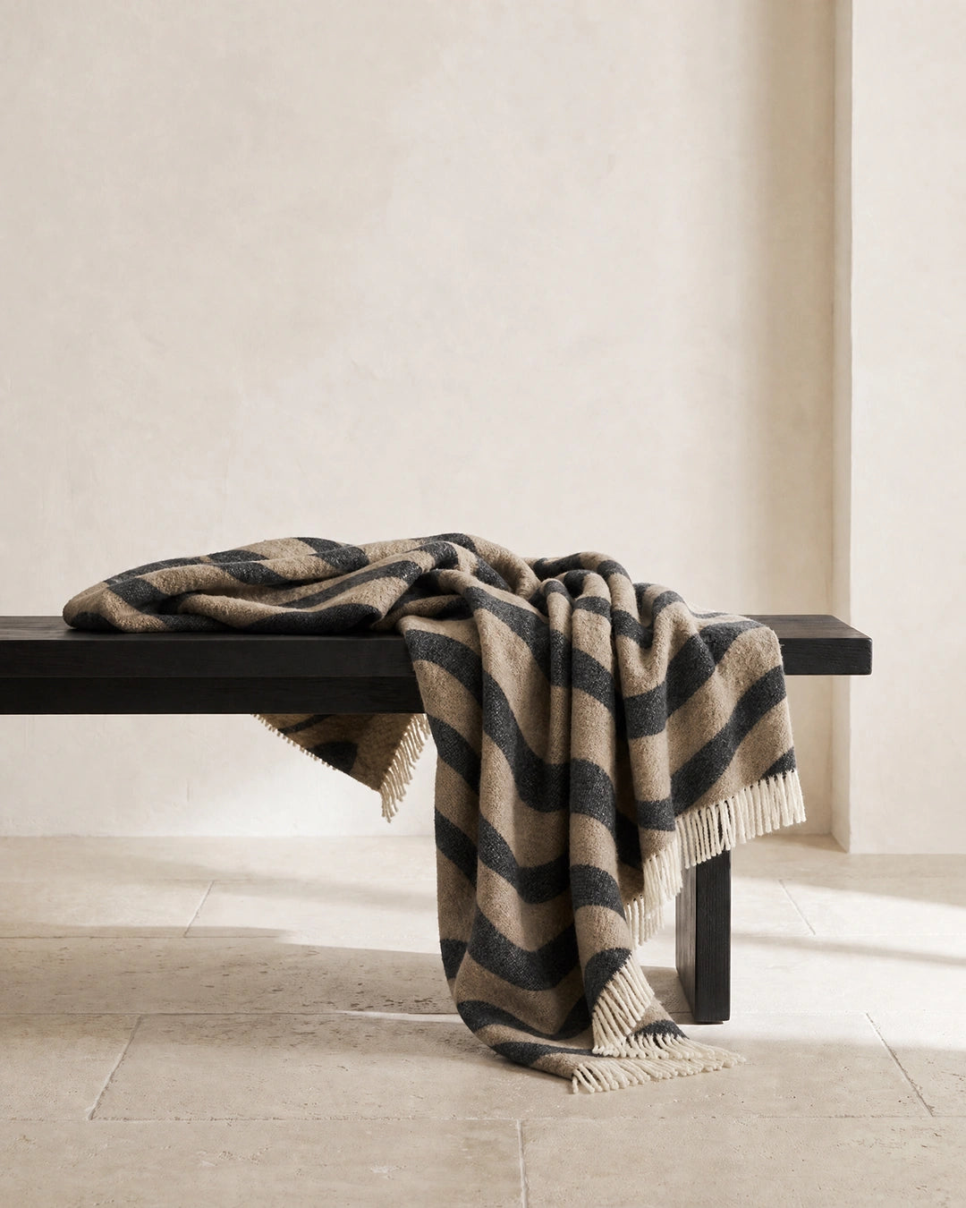 Pläd Flow Beige från Classic Collection i ull med vågformat mönster i svart och beige, draperad över en mörk träbänk.