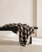 Pläd Flow Beige från Classic Collection i ull med vågformat mönster i svart och beige, draperad över en mörk träbänk.