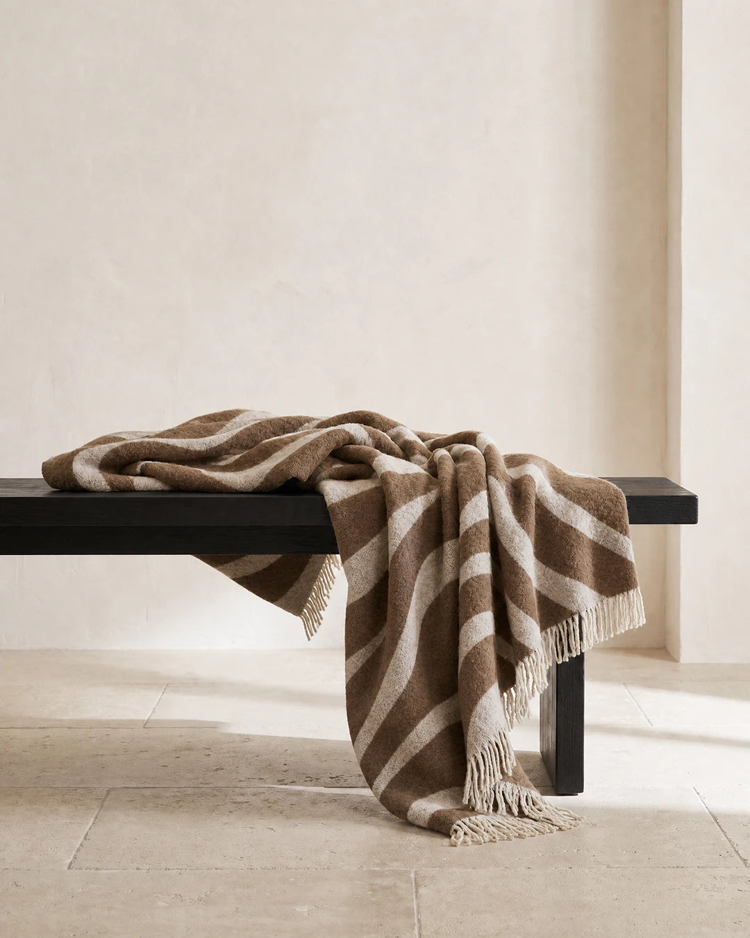 Pläd Flow Brun från Classic Collection i ull med vågformat mönster i brun och off-white, draperad över en mörk träbänk.