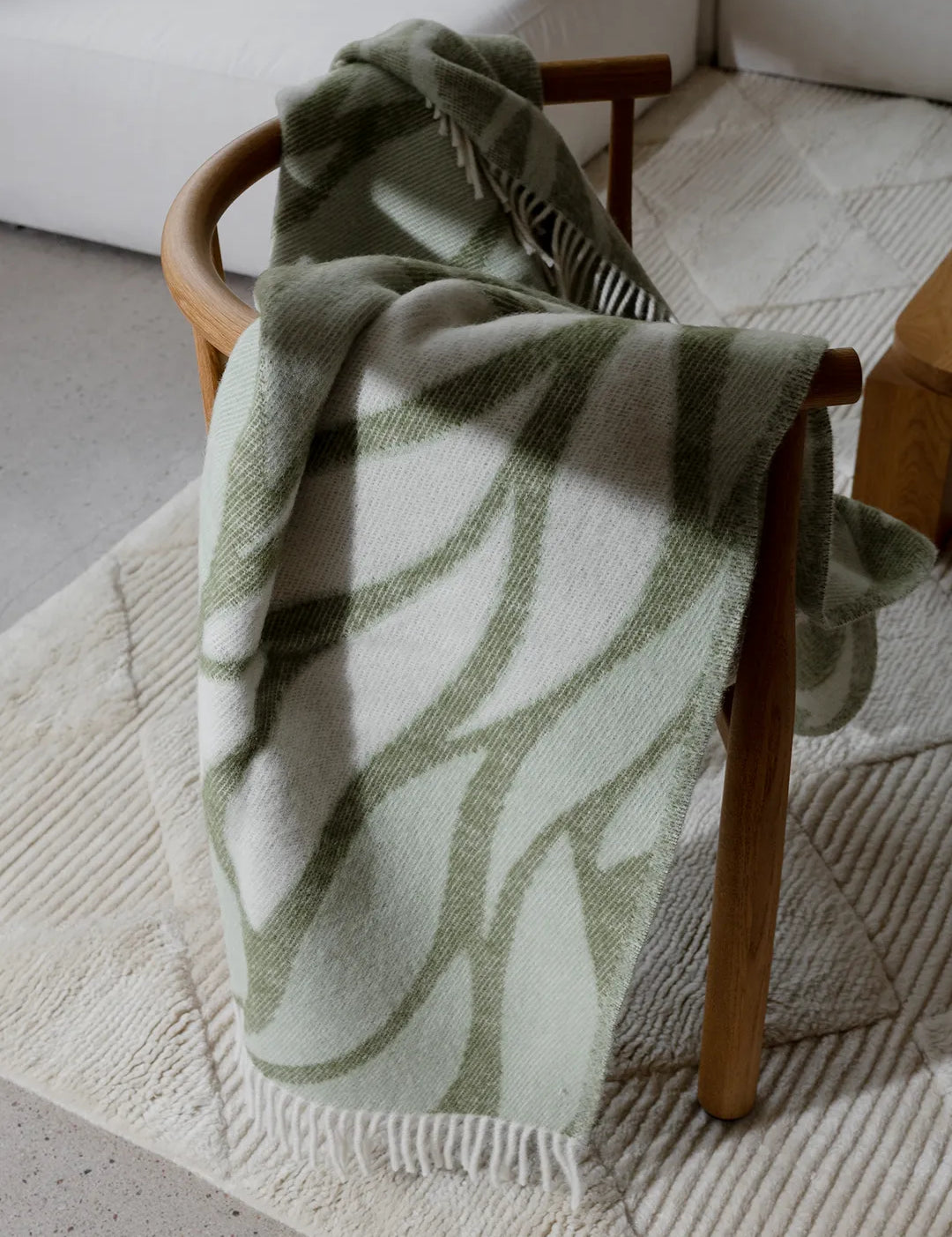 Grönvit pläd Folia i nyzeeländsk ull, draperad över en trästol på en ljus ullmatta.