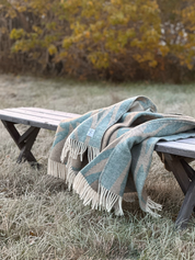 Ullpläd med mönster i gröna och beige toner ligger vikt över en träbänk i frostigt gräs, med höstlöv i bakgrunden.
