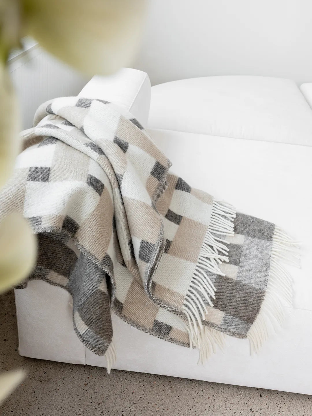 Pläd Weave Beige i nyzeeländsk ull, med rutmönster i beige och grå toner, draperad över vit soffa.