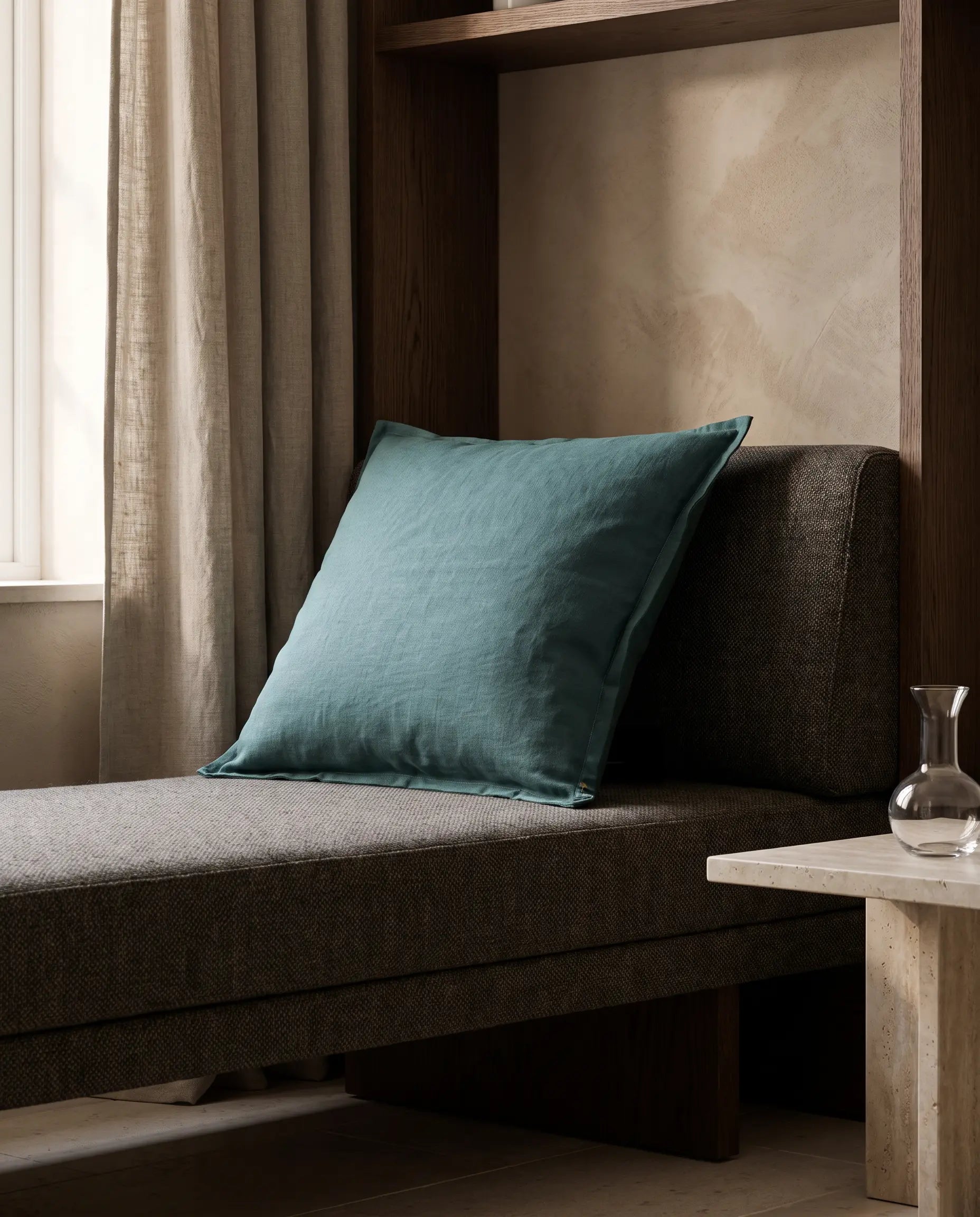 Kuddfodral Classic Linen Teal 50x50 cm från Classic Collection i enfärgat linne, stylat på en mörk stoppad bänk i en varm, elegant interiör.