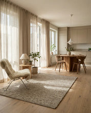Ljus interiör i skandinavisk stil med beige matta, fåtölj i fårskinn, runt träbord och linnegardiner.