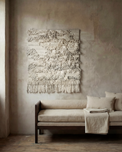 Väggbonad i beige ull med texturerat mönster och fransar, hängande ovanför en minimalistisk beige dagbädd i trä.