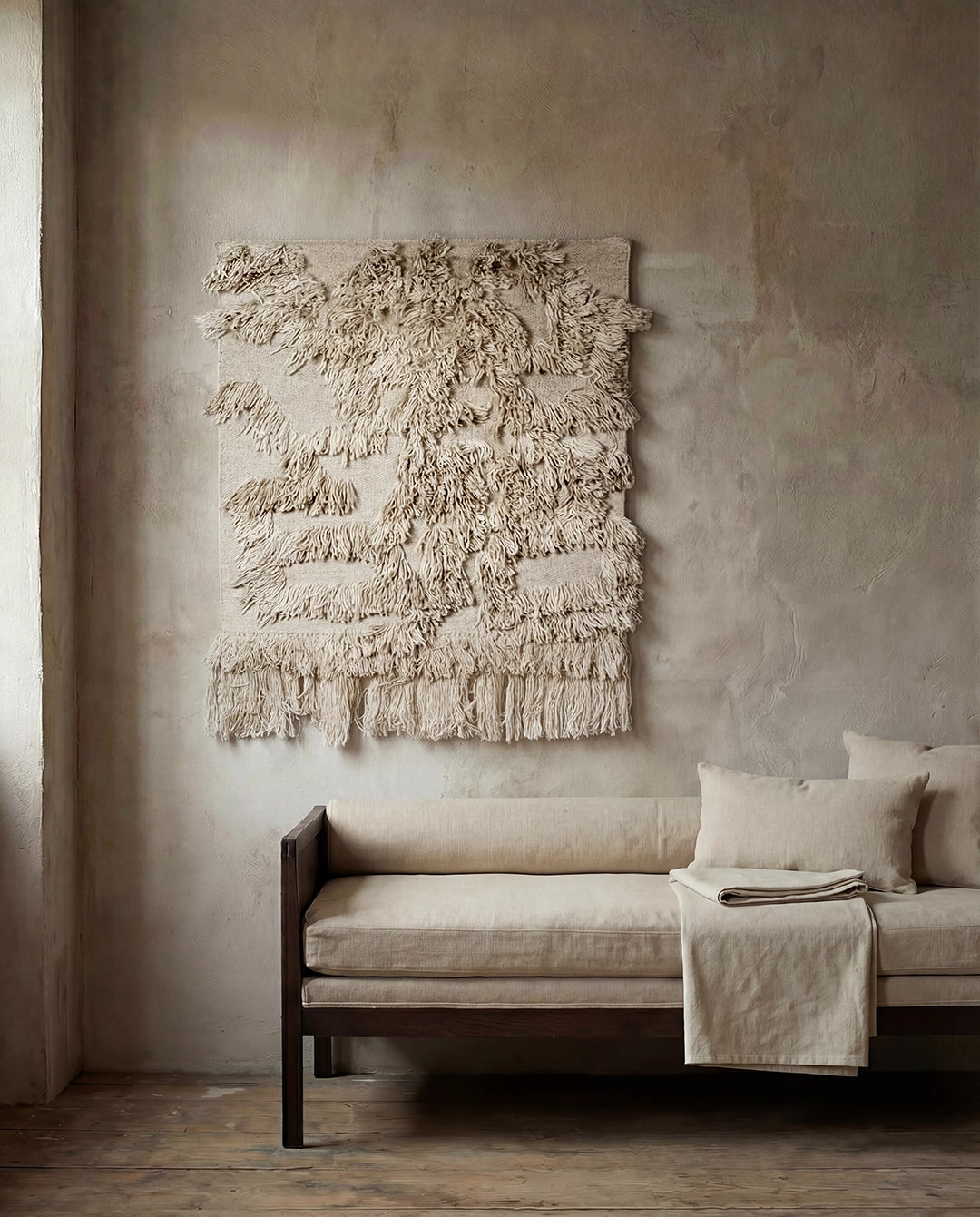 Beige väggbonad i ull med strukturerad textur och fransar, hängande ovanför en minimalistisk beige soffa.