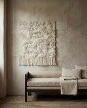 Väggbonad i naturvitt ylle med dekorativ lugg och fransar, hängande ovanför en beige dagbädd i ett stilrent rum.