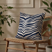 Kudde Zebra Navy, 50x50 cm, med grafiskt zebramönster i mörkblått och naturvitt samt dekorativa fransar längs sidorna, placerad på en flätad trästol framför en beige vägg med klassiska väggpaneler.