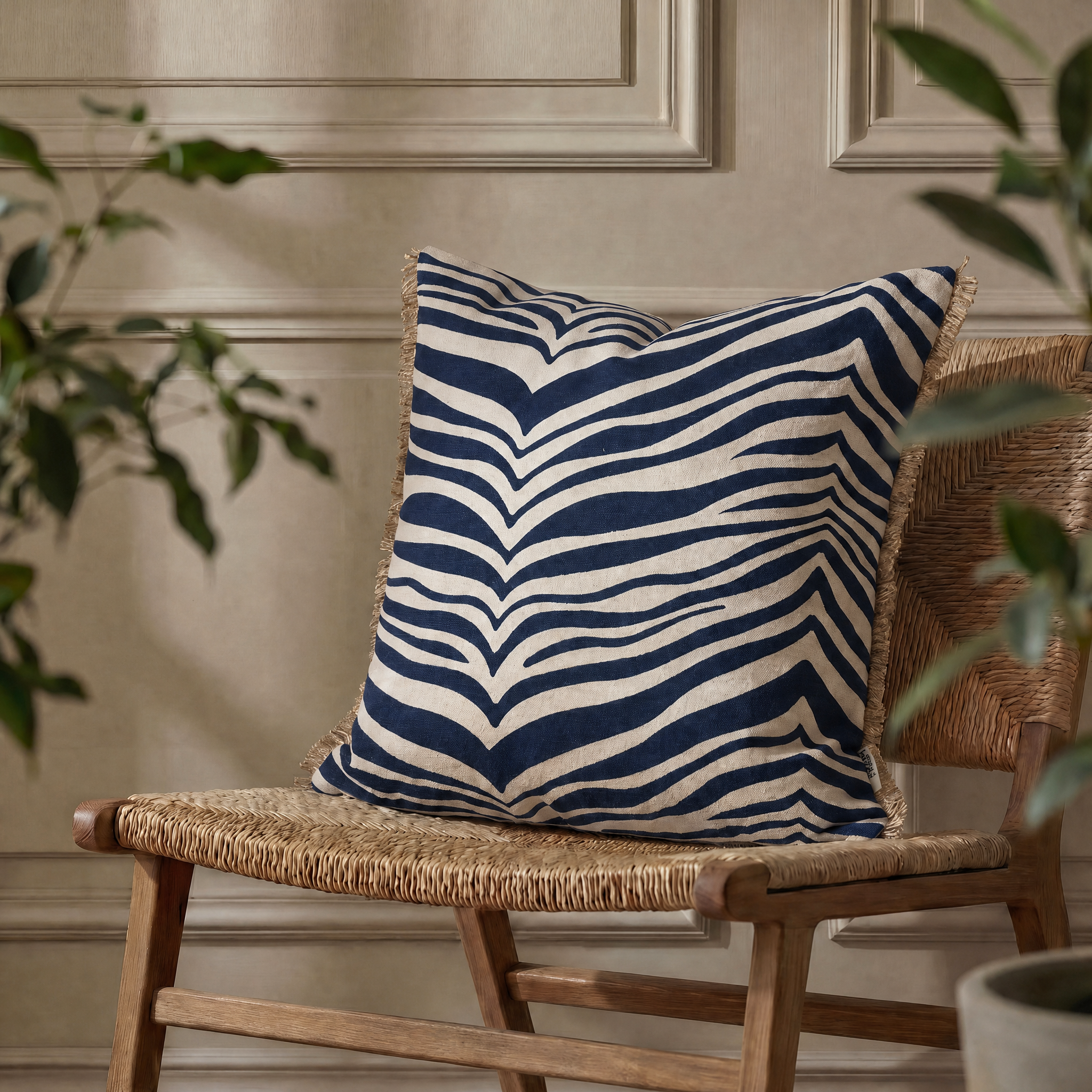 Kudde Zebra Navy, 50x50 cm, med grafiskt zebramönster i mörkblått och naturvitt samt dekorativa fransar längs sidorna, placerad på en flätad trästol framför en beige vägg med klassiska väggpaneler.