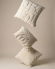 Three floating cream-colored decorative throw pillows with textured geometric and leaf patterns on a beige background.