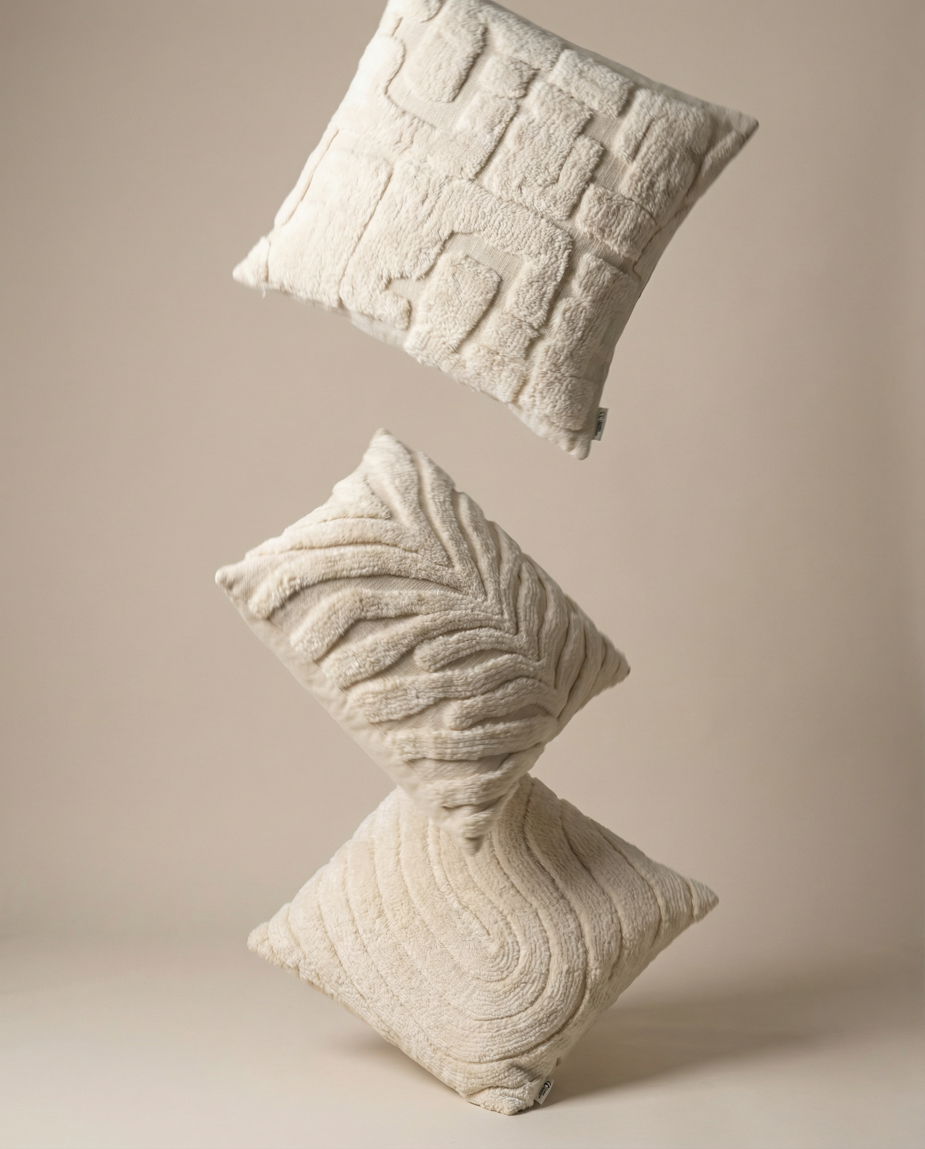 Three floating cream-colored decorative throw pillows with textured geometric and leaf patterns on a beige background.