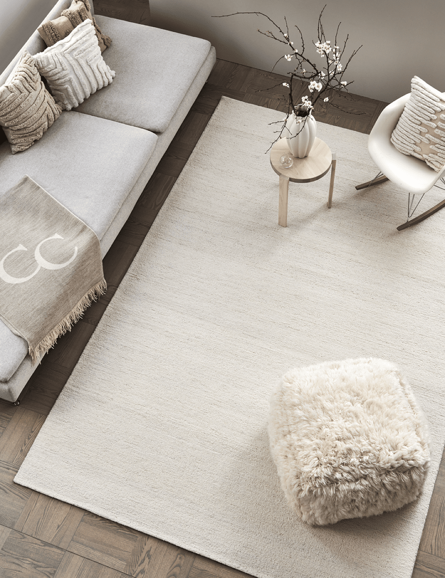 Top-down view of a minimalist beige area rug in a modern living room with a sofa, rocking chair, and faux fur ottoman.