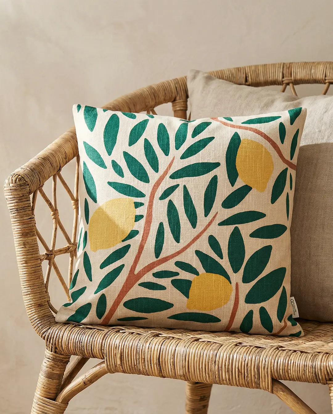 Kuddfodral i linneblandning med mönster av gula citroner och gröna blad på beige botten, placerad i en rottingstol.