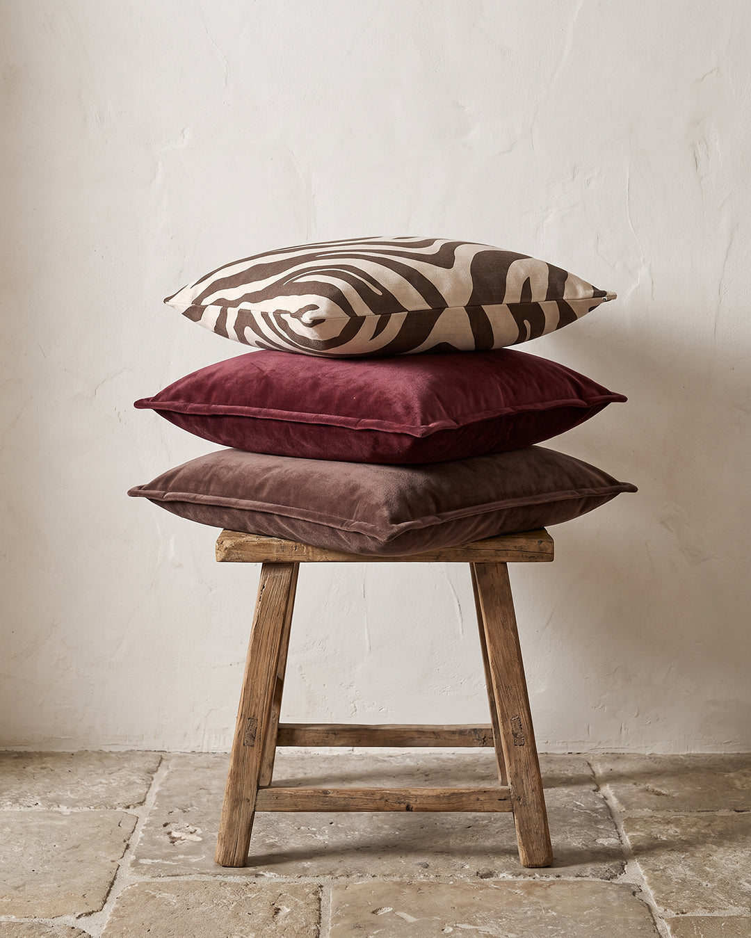 Kuddar 50x50 cm från Classic Collection: Savannah i off-white och brun samt Classic Velvet i burgundy och brun, arrangerade på en träpall med patina.