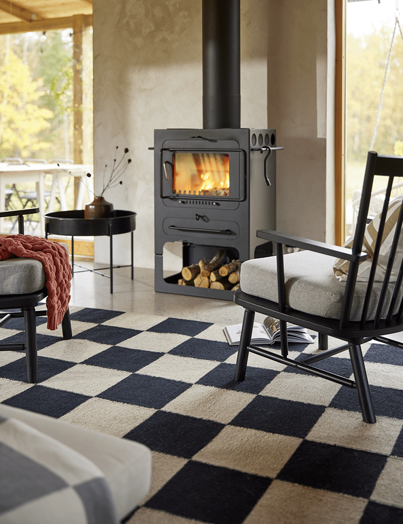 Schwarzer Kaminofen mit brennendem Holz in einem gemütlichen Wohnzimmer mit schwarz-weißem Schachbrett-Teppich.