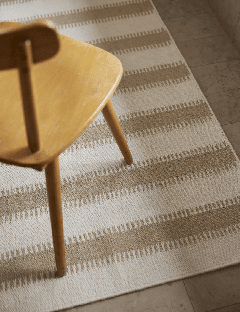 Light beige and cream area rug with textured horizontal stripes under a wooden chair on a tiled floor.