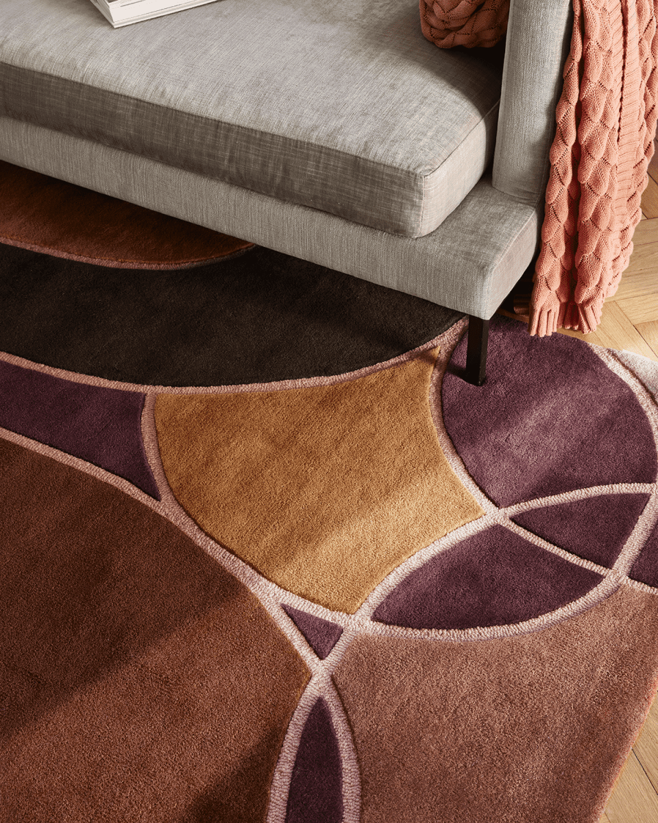 Moderner Teppich mit geometrischem Muster in Brauntönen, Beige und Aubergine vor einem Sofa.