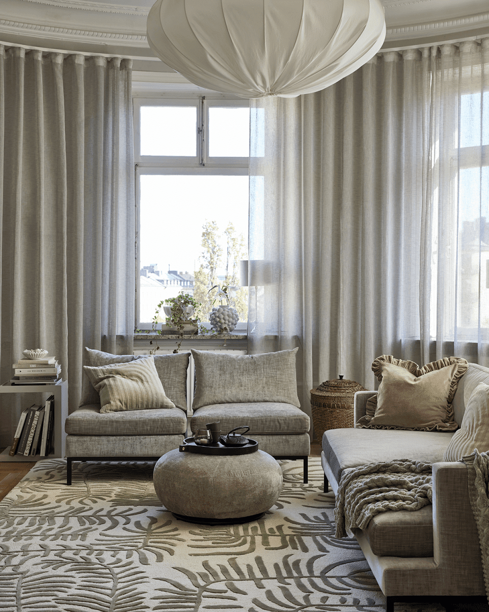 Helles Wohnzimmer mit beige gemustertem Teppich, grauen Loungesesseln, rundem Polsterhocker und eleganten Gardinen.