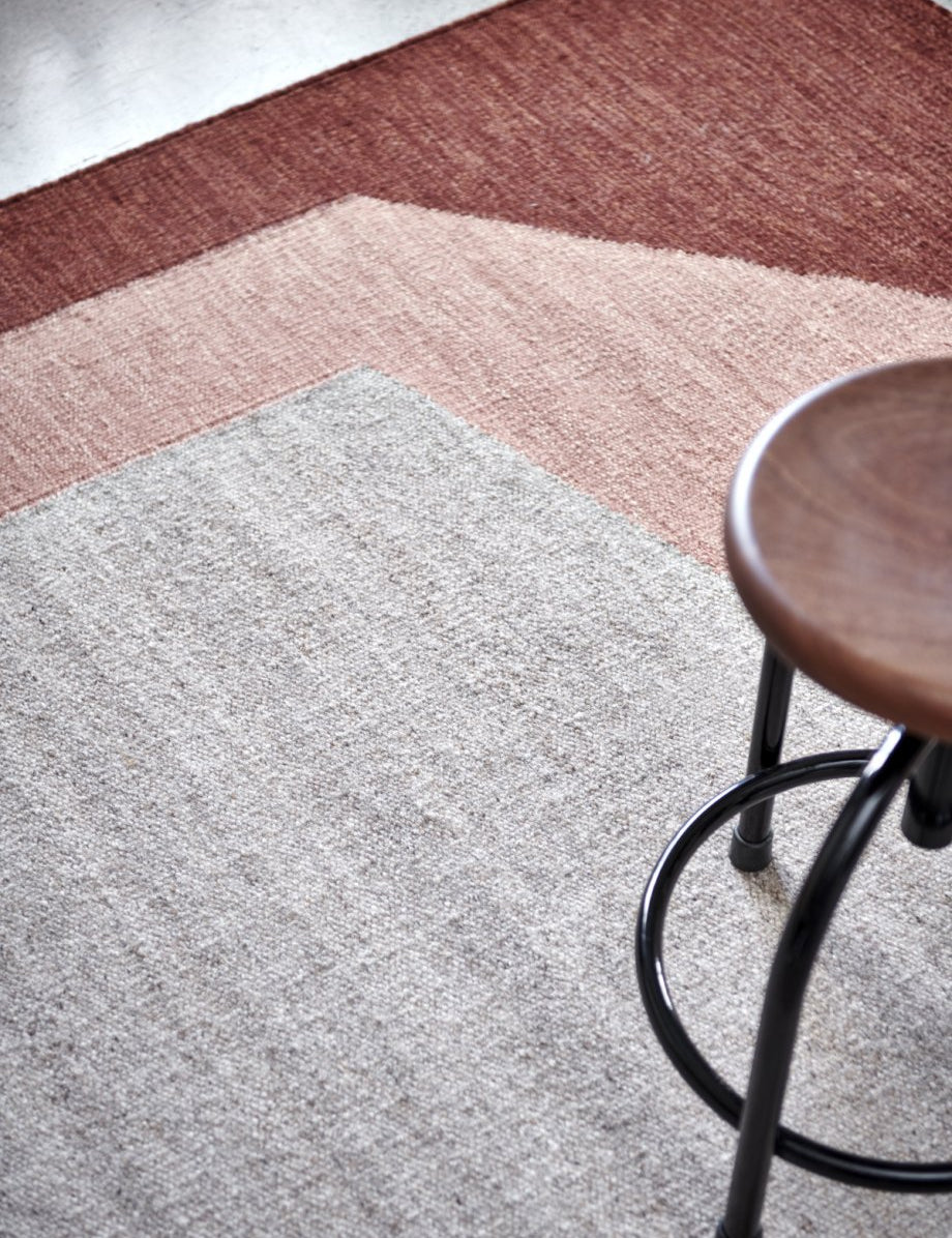 Moderner Webteppich mit geometrischem Farbmuster in Beige, Rosa und Dunkelrot, daneben ein dunkler Holzhocker.